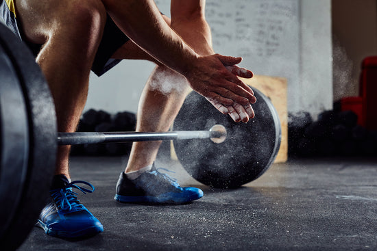 Person in a gym. Barbell on the floor. Person is getting ready to deadlift the weight off the floor. They are squatting down and rubbing chalk in their hands.